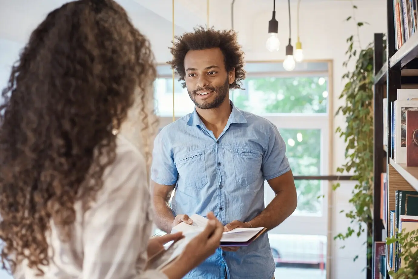 Крупный план двух молодых людей у книжной полки в библиотеке: они держат книги и обсуждают студенческую жизнь. Привлекательный темнокожий студент разговаривает с симпатичной девушкой об экзаменах.